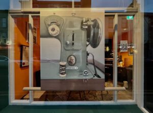 A large realistic painting of a vintage sewing machine is displayed in a window, partially obscuring the view of a warmly lit interior with orange walls and framed items. Reflections of buildings are visible in the window glass.