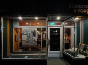 A storefront at night displays a vintage telephone mural in the left window, a black door with signage in the center, and an old typewriter in the right window. The interior is warmly lit.