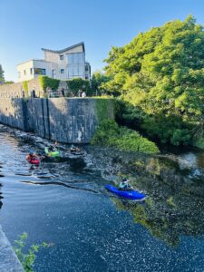 Four people kayak along a narrow river bordered by a stone wall and lush green trees, with a modern building and people standing above on the left. The sky is clear and blue.