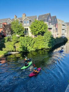 Four people kayak on a calm river bordered by lush greenery and colorful houses under a clear blue sky. The sunlight casts bright reflections on the water.