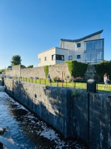 A modern house with large windows stands behind a stone wall next to a canal, with people sitting and walking along the grassy path beside the water under a clear blue sky.