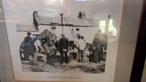 A black-and-white photo shows people gathered around a tripod crane at a village market in front of old cottages. Several figures carry baskets or sacks. Caption: The Small Crane potato and turf market.