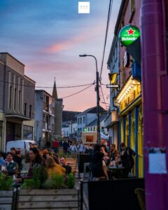 Outdoor street scene at sunset with people dining and socializing at tables outside colorful restaurants and pubs. The street is lively, and a Heineken sign glows above one of the buildings.