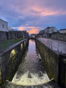 A narrow canal with stone walls runs through a town at sunset, reflecting colorful clouds in the sky. Buildings line both sides, and soft lights illuminate the canal’s edges.