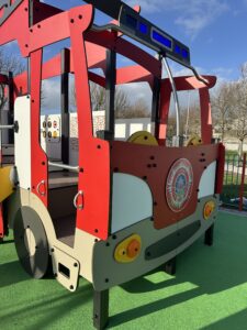 A colorful playground structure shaped like a fire truck sits on green rubber safety flooring under a blue sky with some clouds and bare trees in the background.