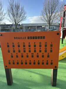 An orange playground sign displays the Braille alphabet and numbers, with black printed letters, numbers, and Braille symbols. The sign stands on a green surface with trees and buildings in the background.