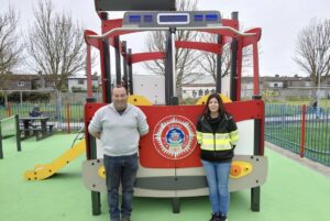 Two adults stand in front of a fire truck-themed playground structure with a slide, on green safety flooring. Leafless trees and houses are visible in the background behind a blue fence.